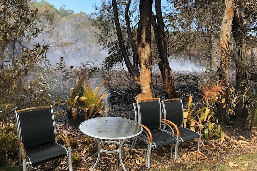 Burnt and grass and trees behind a backyard. Singed table and chairs sit at the edge of the backyard.
