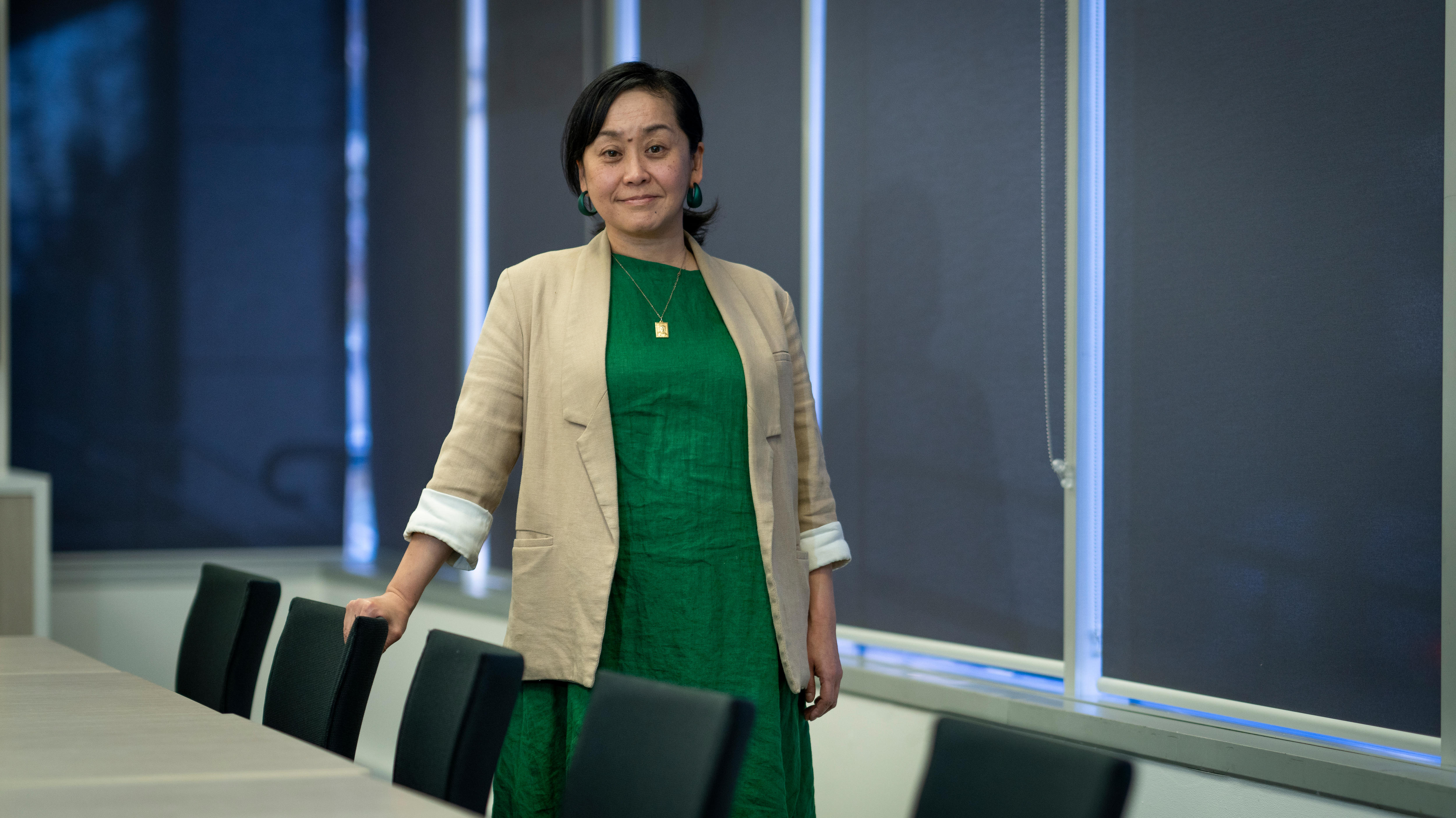 A woman in a green dress and white blazer stand behind a desk