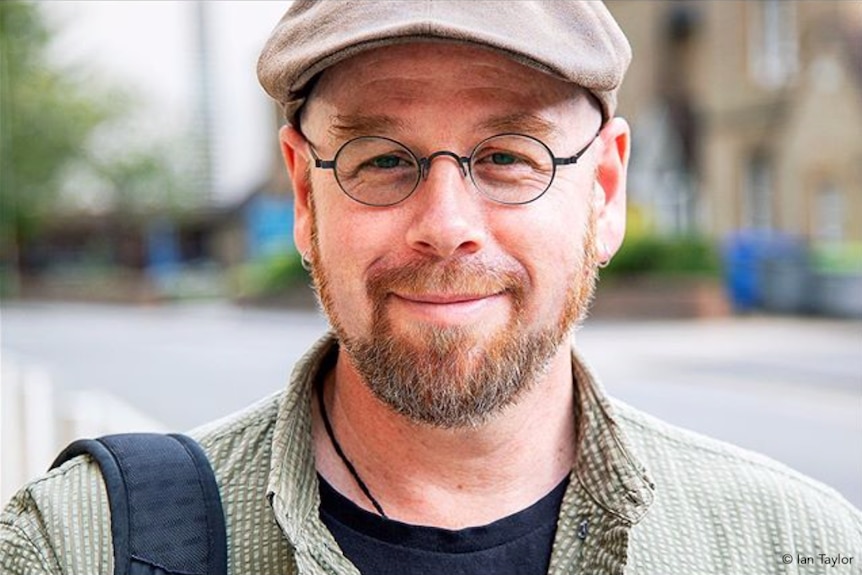 Un hombre lleva gafas con un sombrero plano de tweed y sonríe.