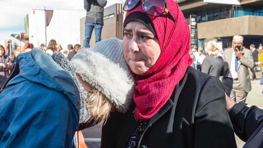 A woman in a blue jacket leans against a woman wearing a red head scarf.