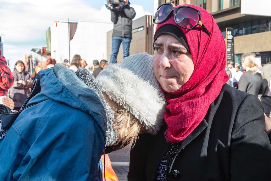 A woman in a blue jacket leans against a woman wearing a red head scarf.