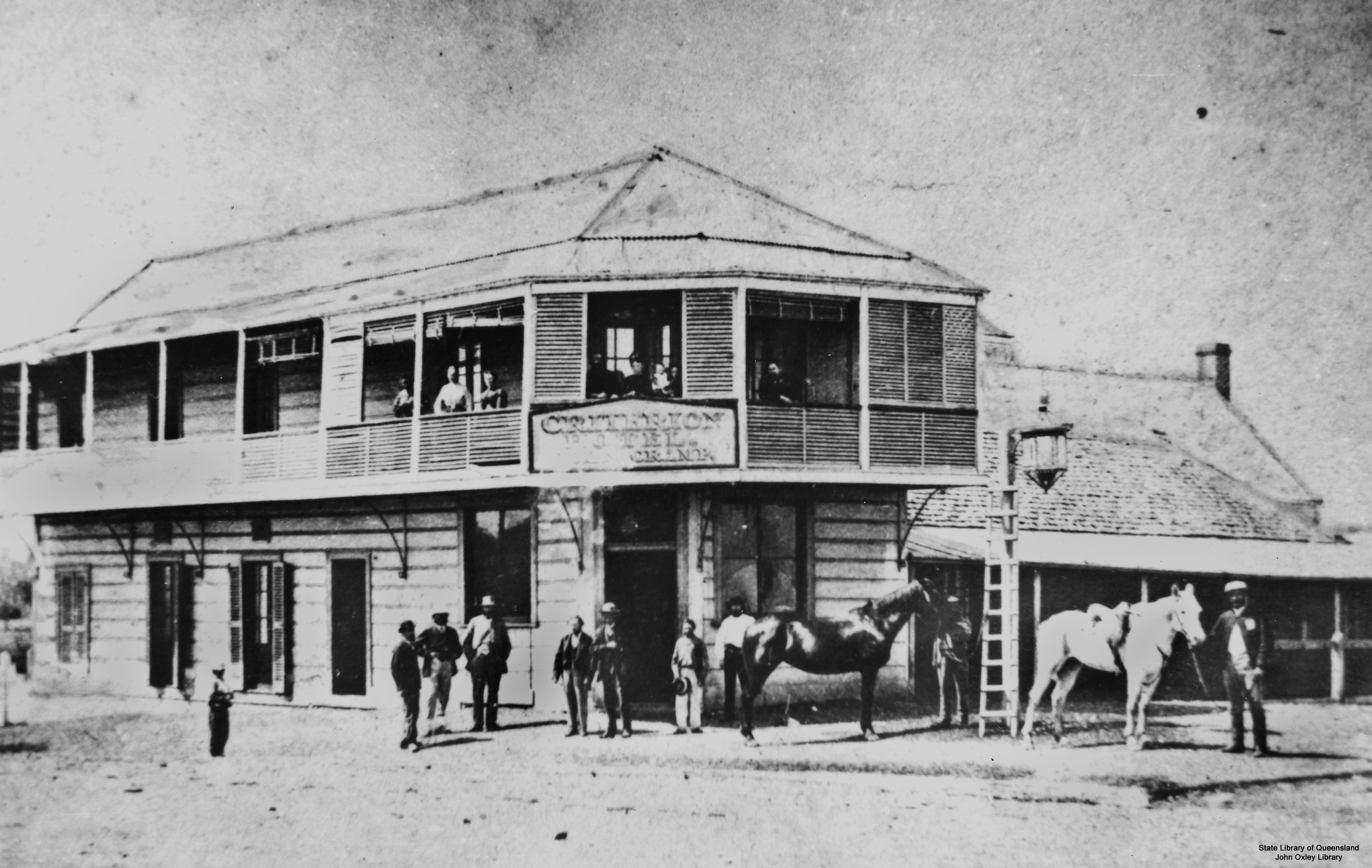 Black and white photo of two storey timber hotel, men, horses standing around