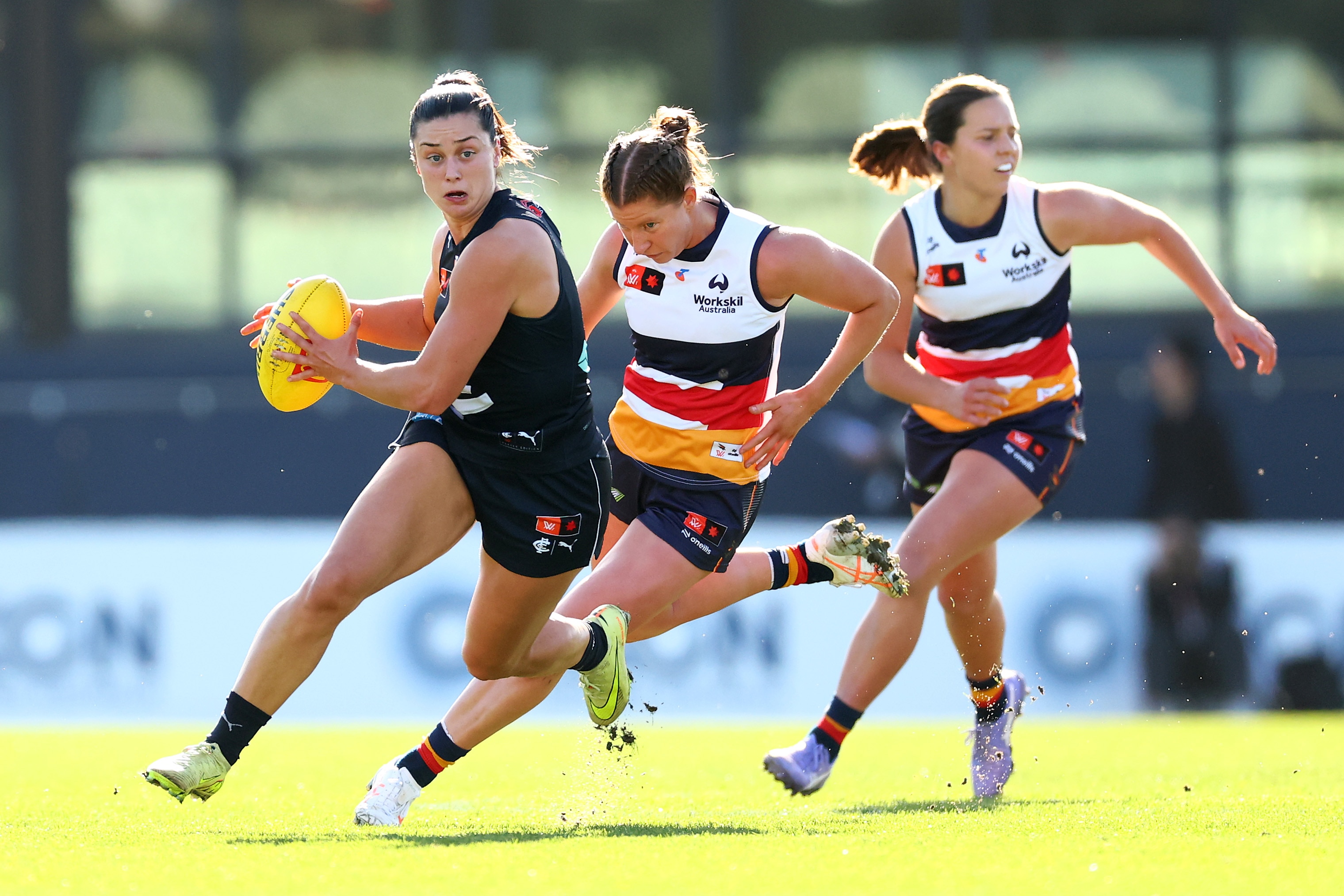 A Carlton AFLW player runs with the ball while being chased by two Adelaide defenders.