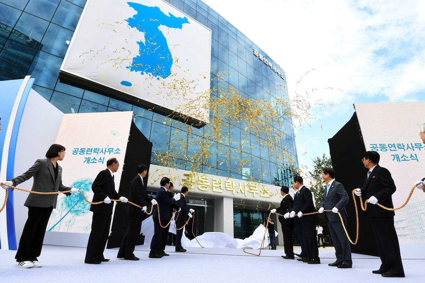 Two lines of suited officials tug a golden rope while wearing white gloves as part of an opening ceremony for a glass building.