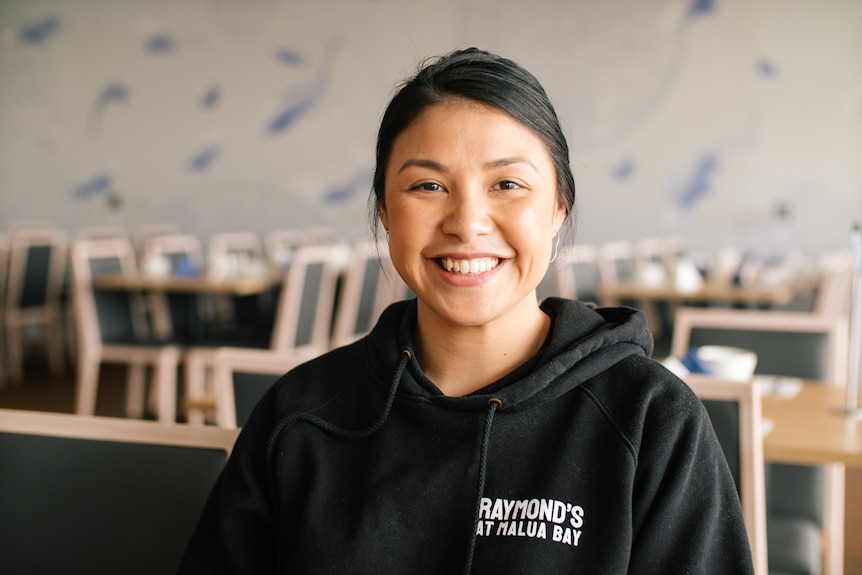 A young woman with black pulled back hair smiles as she sits at an table in an empty restaurant. 