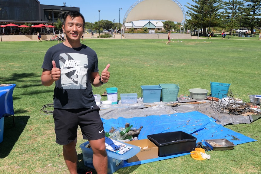 Man smiles with his thumbs at park in front of collection of rubbish found by him and fellow divers 