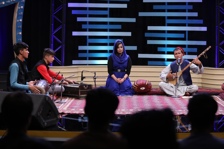 A man plays a musical instrument on TV while a woman in a headscarf watches. 