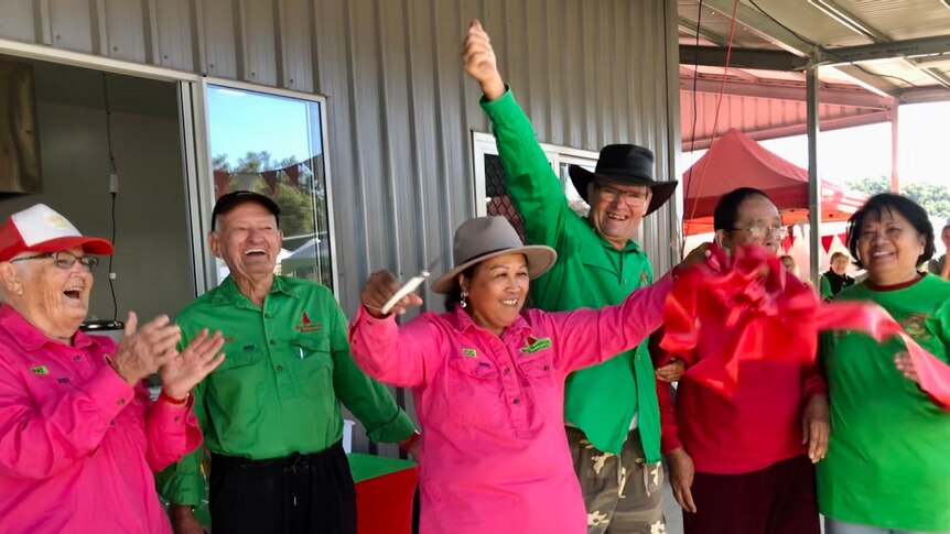 Six people celebrate after cutting a ribbon to the new shed behind them.