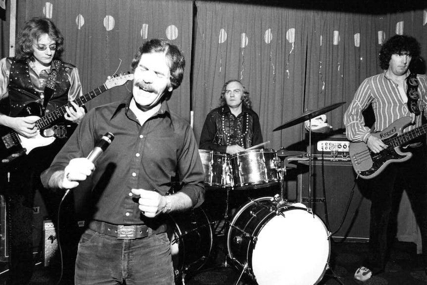 A four-piece rock band, fronted by a moustached singer, perform in this black and white photo.