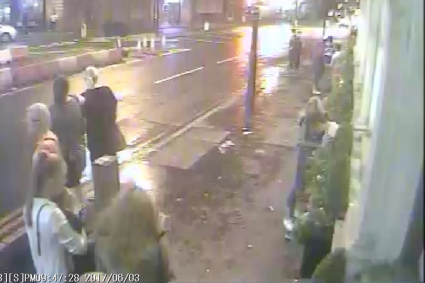 Two women walk with their backs to the camera as other people walk in the street ahead of them.