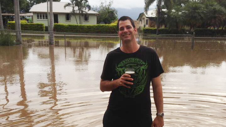 Giru resident Richard Berryman stands holding a beer in floodwaters outside his house.