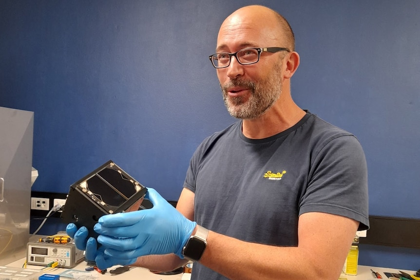 a bald-headed man with a grey beard holds a tiny satellite, measuring about 10 centimetres along all sides