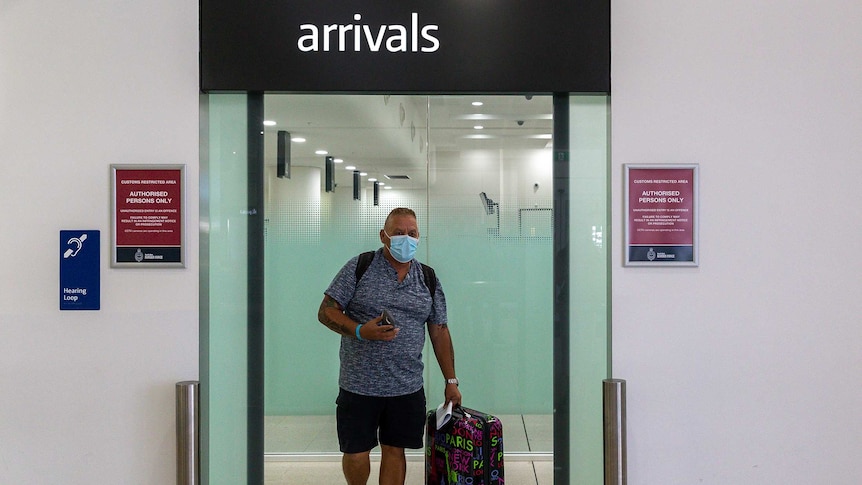 A man wearing a facemask and pulling a suitcase walks through the arrival gates inside an airport terminal.