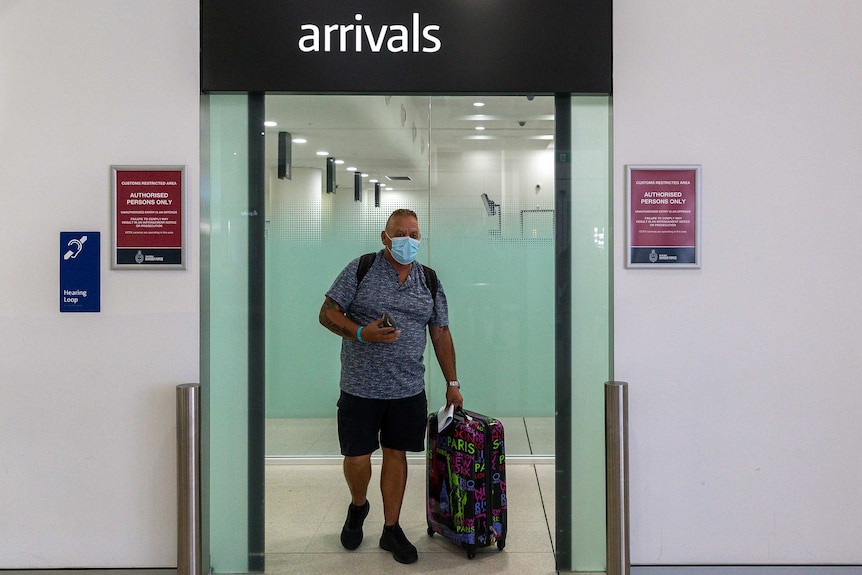 Un homme portant un masque facial et tirant une valise traverse les portes d'arrivée à l'intérieur d'un terminal d'aéroport.