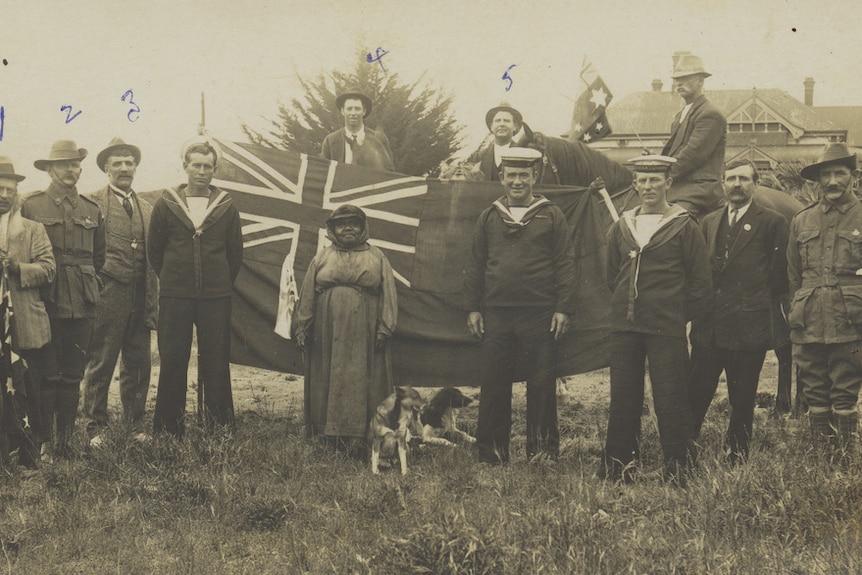 black and white photo of soldiers in front of an Australian flag with a smiling Indigenous woman centre