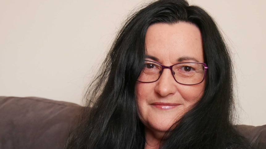 A head and shoulders image of a woman with long black hair and purple glasses sitting on a couch.