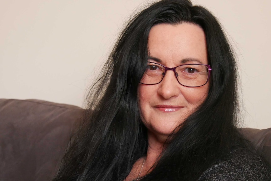 A head and shoulders image of a woman with long black hair and purple glasses sitting on a couch.