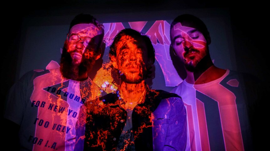 From left to right, Paul Bender, Simon Mavin and Perrin Moss stand together with a colourful, abstract image projected on them.