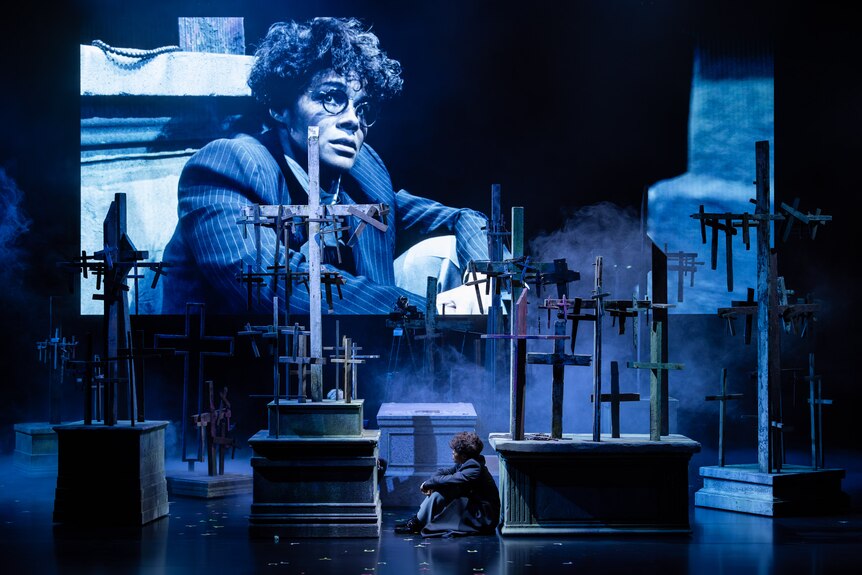 On stage, Zahra Newman sits in a graveyard. Behind her is a screen, showing her in a man's wig and glasses, in close-up.