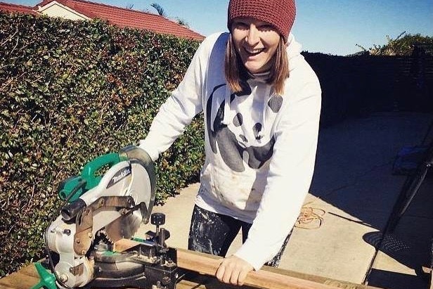 A woman stands at an outdoor workbench holding a drop saw machine, smiling.