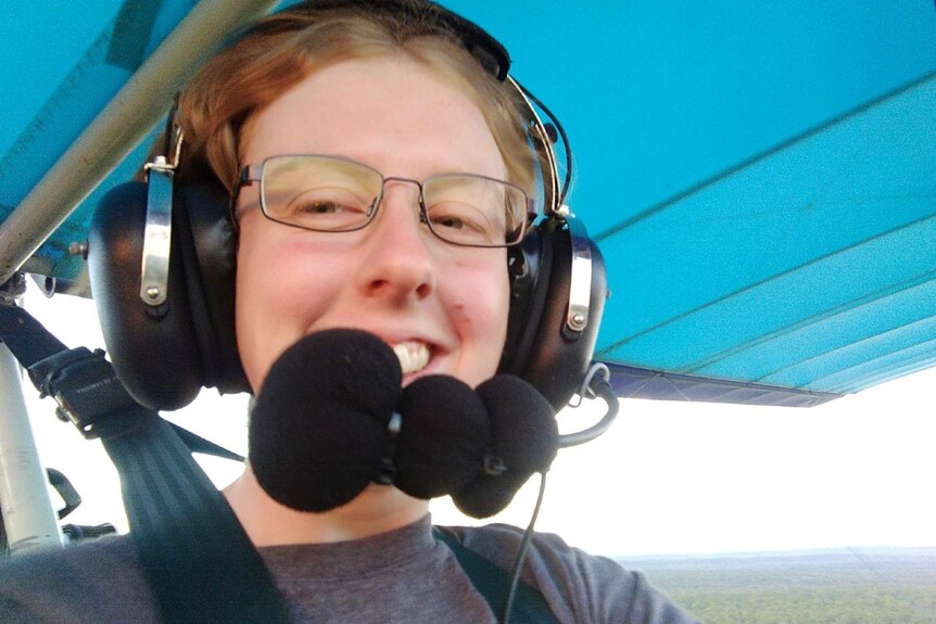 A woman in glasses and a pilot headset smiles from inside an open-air cockpit on a sunny day.