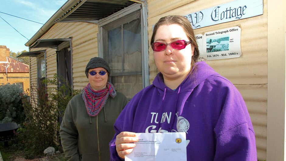 Two women, one holding a NPWS notice outside an old country cottage