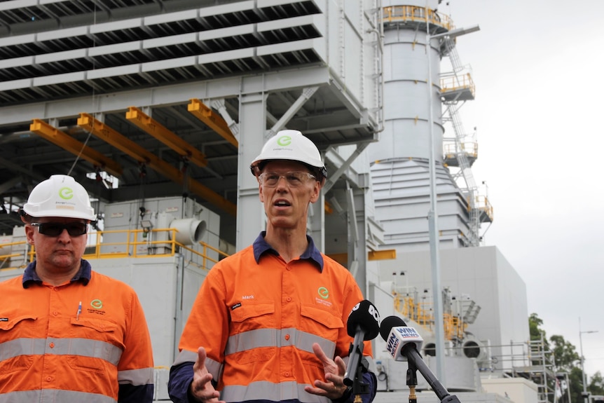 Two men in high-vis and safety wear stand in front of industrial power plant