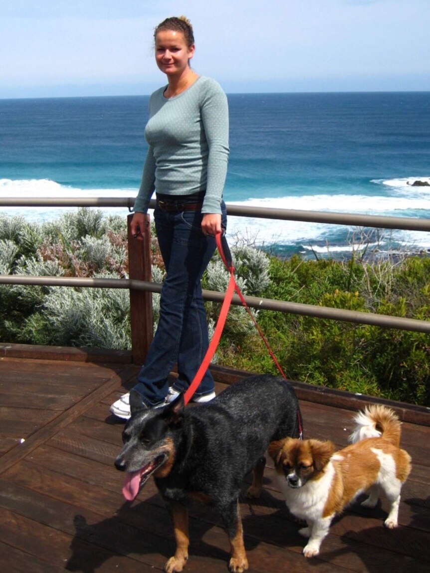 Roxy stands on a wooden path near the ocean. She is smiling and holding the leads of two dogs.