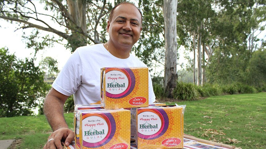 An Indian man kneeling behind a box of coloured packets of power in a green, leafy park.