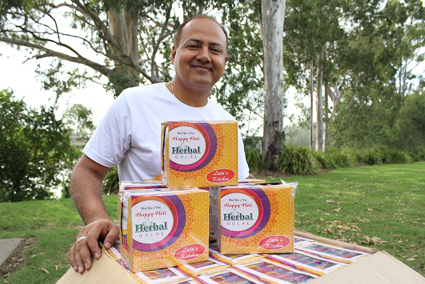 An Indian man kneeling behind a box of coloured packets of power in a green, leafy park.
