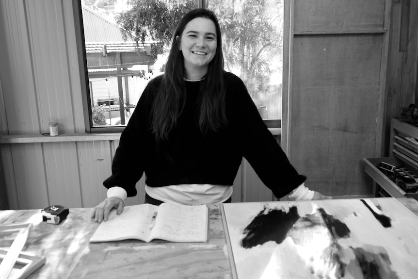 A black and white photo of a woman smiling at the camera with a tape measure, art and timber on the table