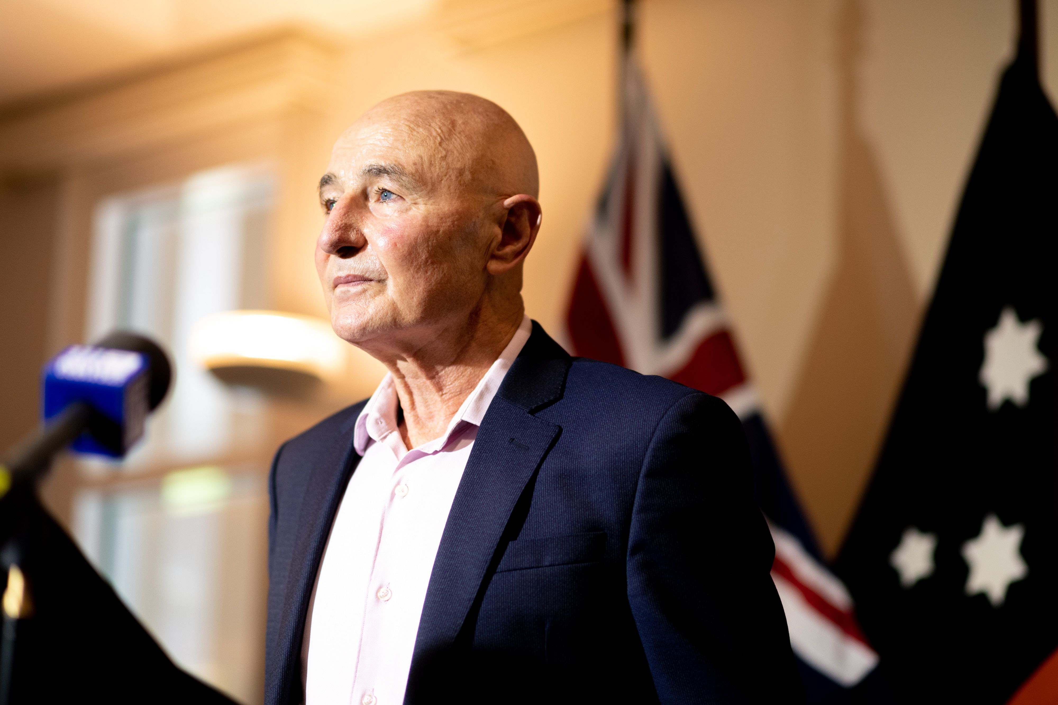 A man wearing a suit stands in front of a microphone with a serious expression.