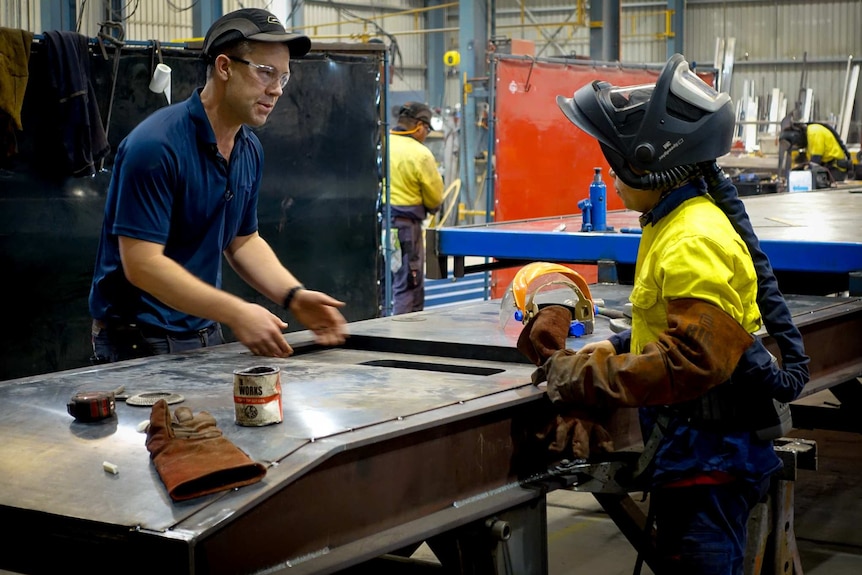 Jason stands at a welding bench talking to an employee.