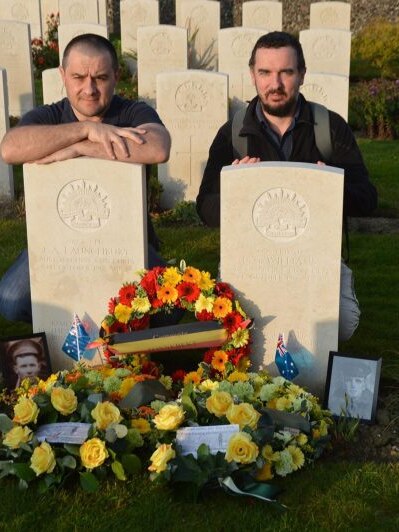 Two men wearing dark shirts, pose behind a photo two graves surrounded by photos and flowers