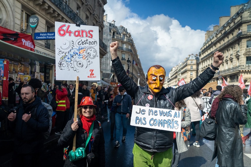 In the middle of a protest a man wears a crude mask of Macron's face, and a woman holds a sign depicting an old mechanic