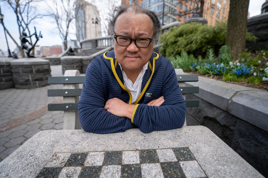 Man with his arms crossed sits at a chess table in a New York park.