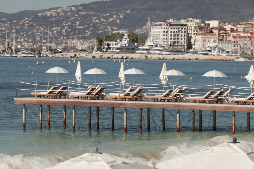 Sunbeds are lined up on a pier in  Cannes, France. There are many ships in the harbor, and lots of hi-rise buildings.