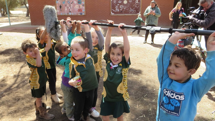 Cameraman filming children hanging off boom pole with microphone on end in school playground.