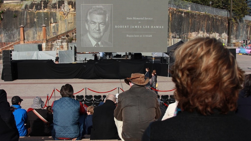 People sit on steps looking down on a stage on which a large sign has been erected for a memorial.