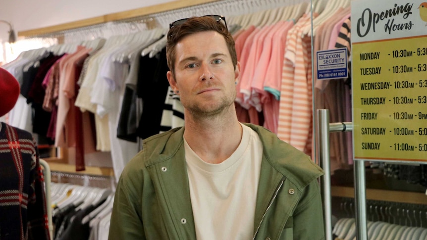 A man standing in a shop with racks of clothes on the wall behind him.