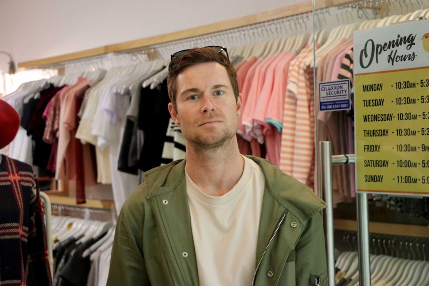 A man standing in a shop with racks of clothes on the wall behind him.