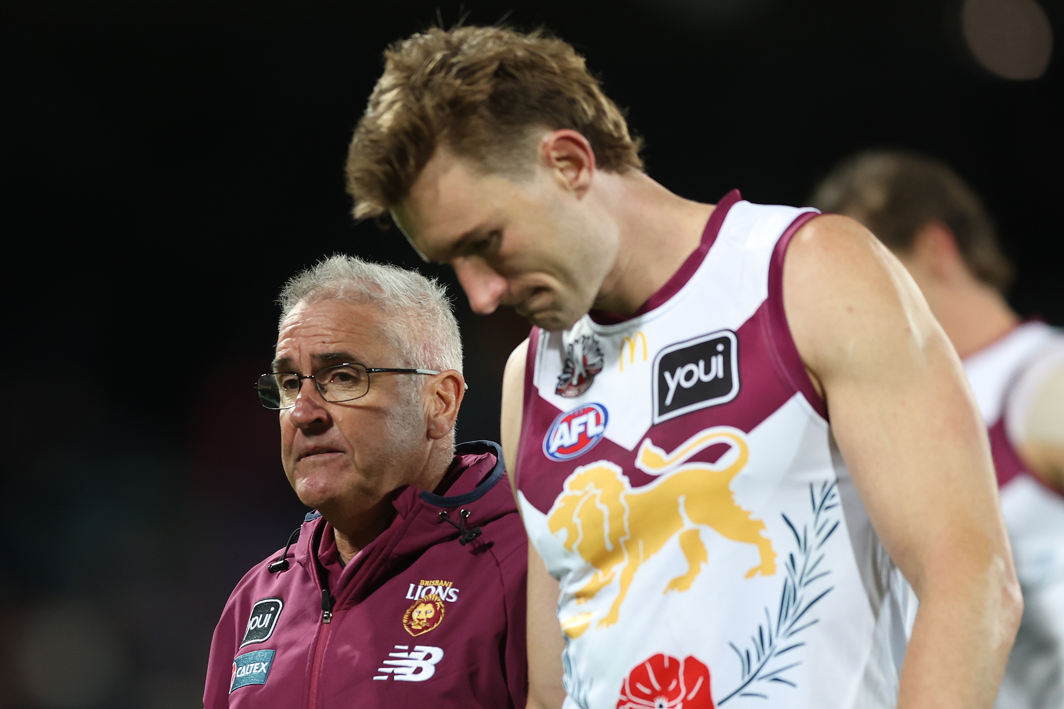 Um técnico do Brisbane AFL de aparência triste caminha enquanto o capitão do Lions caminha com ele de cabeça baixa.