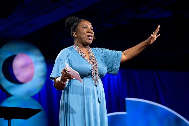Tarana Burke, wearing a long light blue dress, speaks on stage at a TED conference.