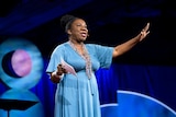 Tarana Burke, wearing a long light blue dress, speaks on stage at a TED conference.