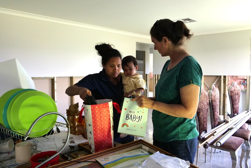 A woman carrying a baby talks to another woman and they rummage through gift bags 