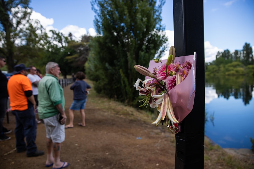 A bunch of flowers is taped to a pole on the riverbank. Some people stand solemnly, looking out onto the water.