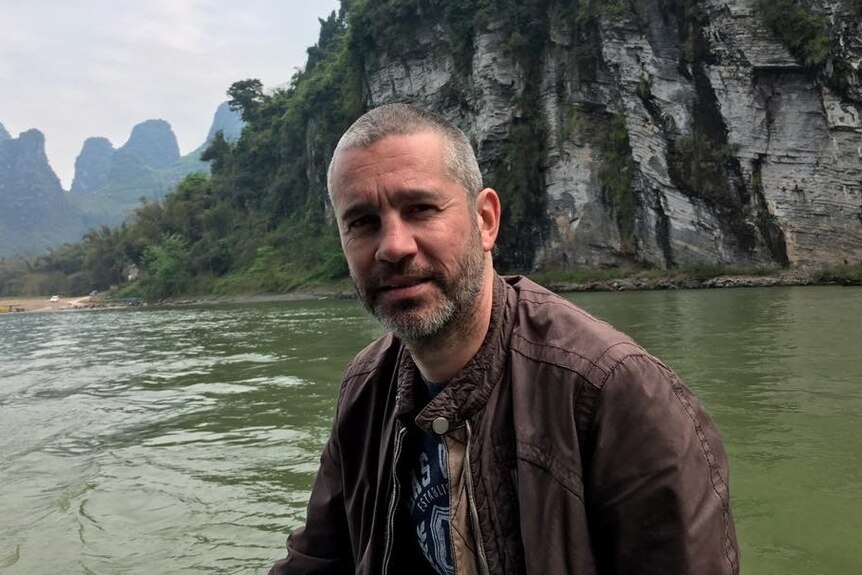 A man with short grey hair and a beard sitting on a boat with a cliff face and other mountains in the background.
