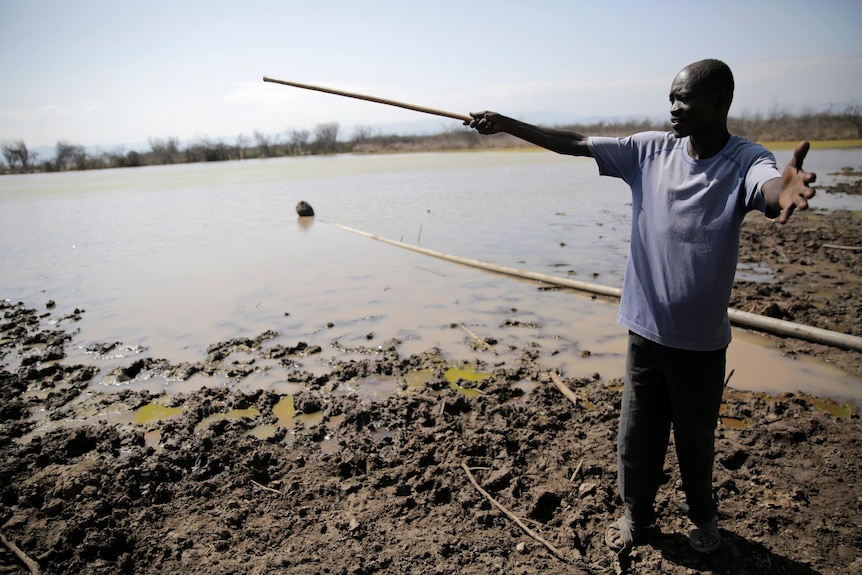 Laban Keben holds his arms wide, holding a stick, at the lake shore 