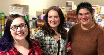 Three women stand with food items stacked in crates and on desks.