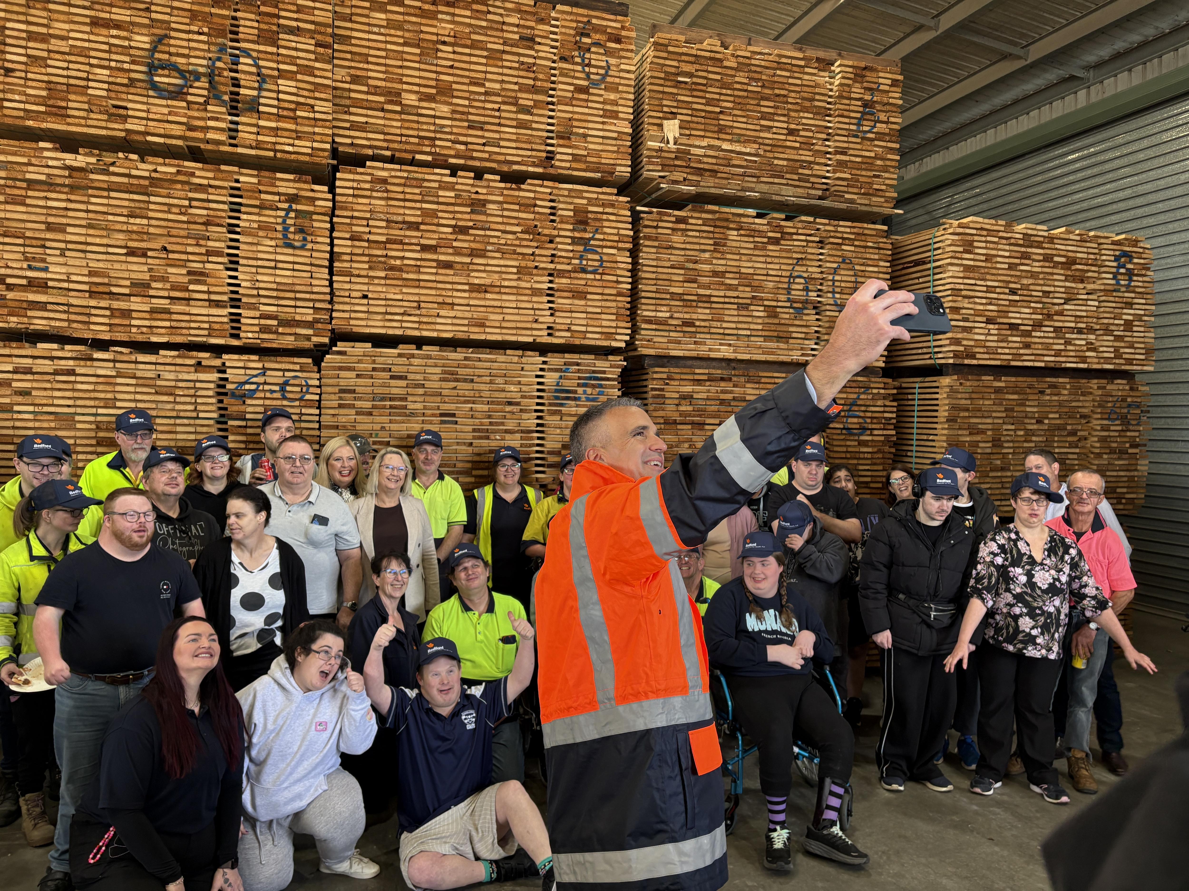 A man in an orange vest holding a phone in front of a crowd with timber in background